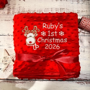 May include: A bright red dimpled minky baby blanket featuring an embroidered reindeer with gold antlers and a red nose. The text reads Ruby's 1st Christmas 2026 with white snowflakes. A red satin ribbon is tied around the center of the blanket.