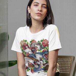 Op de afbeelding: Wit T-shirt met een kleurrijk grafisch ontwerp van slangen en rozen. Het T-shirt met korte mouwen wordt gecombineerd met een zwarte broek en witte sneakers. Het model zit op een houten stoel in een kamer met planten.