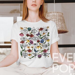 May include: A white t-shirt featuring a colourful print of eyes, flowers, and butterflies. The print is a whimsical and abstract design. The t-shirt is being worn by a person sitting in a yoga pose.