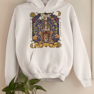 May include: White hoodie with a graphic print of a lion and a woman with a crown of leaves. The graphic is surrounded by stars, moons, and a zodiac wheel. The text "Leo" and "&Omega; LEO &Omega;" is printed on the graphic.
