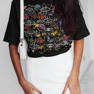 May include: Black t-shirt with a colourful floral print featuring stylised eyes, butterflies, and celestial symbols.