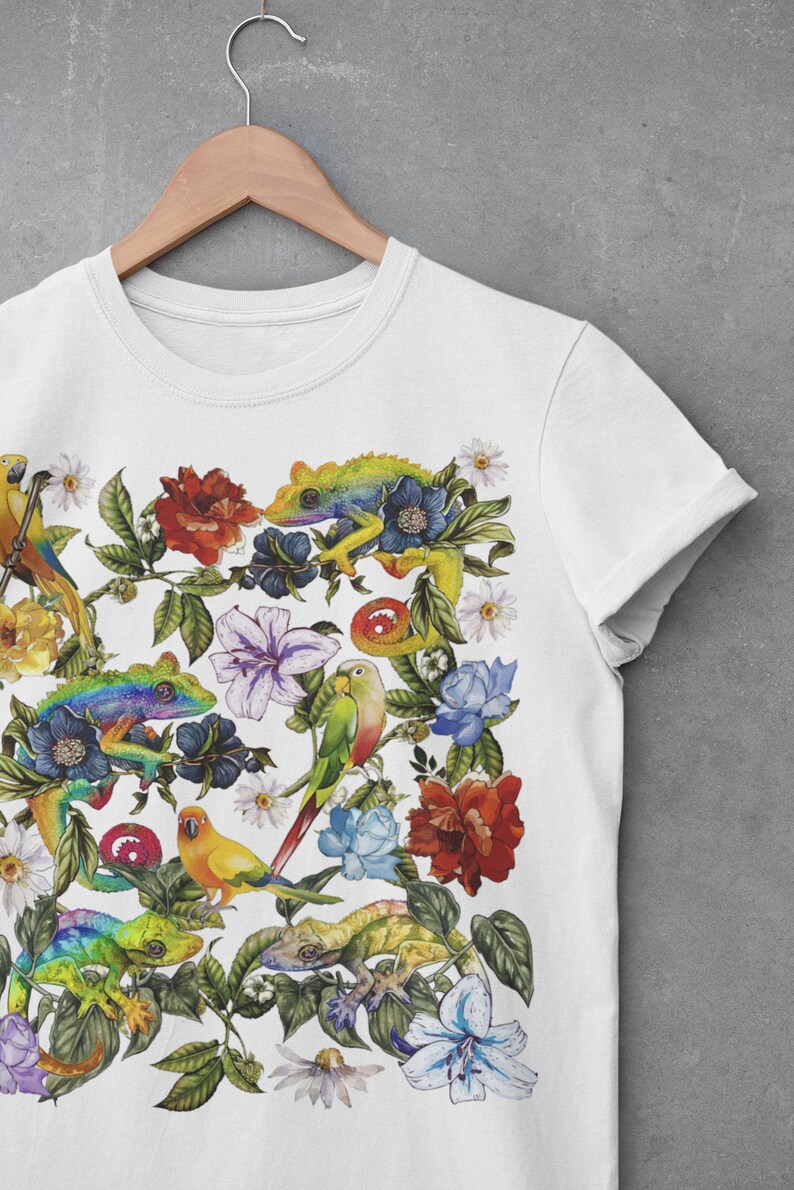May include: White t-shirt with a colorful print of parrots, lizards, and flowers. The design features a variety of tropical plants and animals in a vibrant, whimsical style.