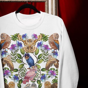 May include: White sweatshirt featuring a colorful owl and floral design. The design includes various owls in shades of brown, blue, and pink, surrounded by blue and purple flowers and green leaves. The sweatshirt is hanging on a black hanger.