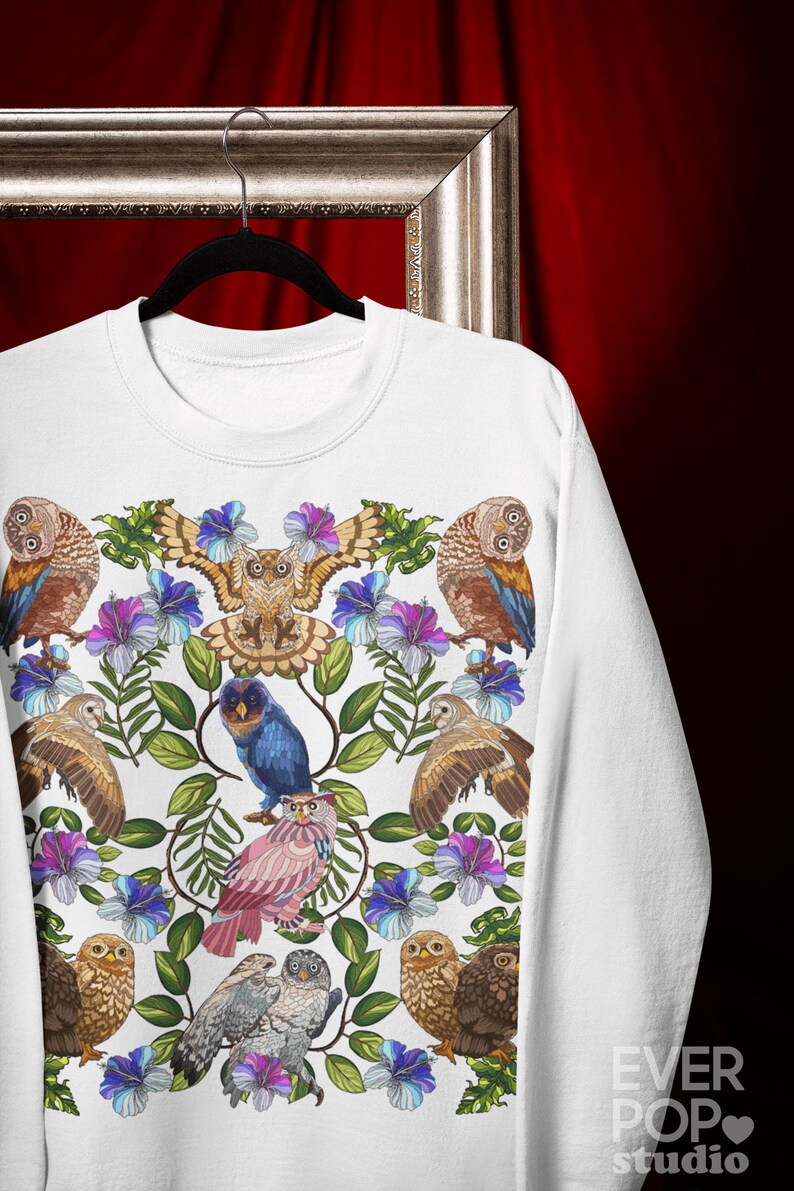 May include: White sweatshirt featuring a colorful owl and floral design. The design includes various owls in shades of brown, blue, and pink, surrounded by blue and purple flowers and green leaves. The sweatshirt is hanging on a black hanger.