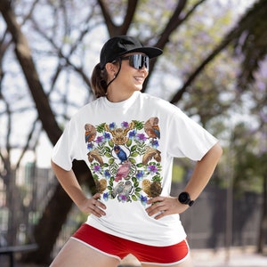 May include: White t-shirt featuring a colorful bird and floral design. The shirt has a crew neck and short sleeves. The person is wearing red shorts and a black cap and sunglasses.