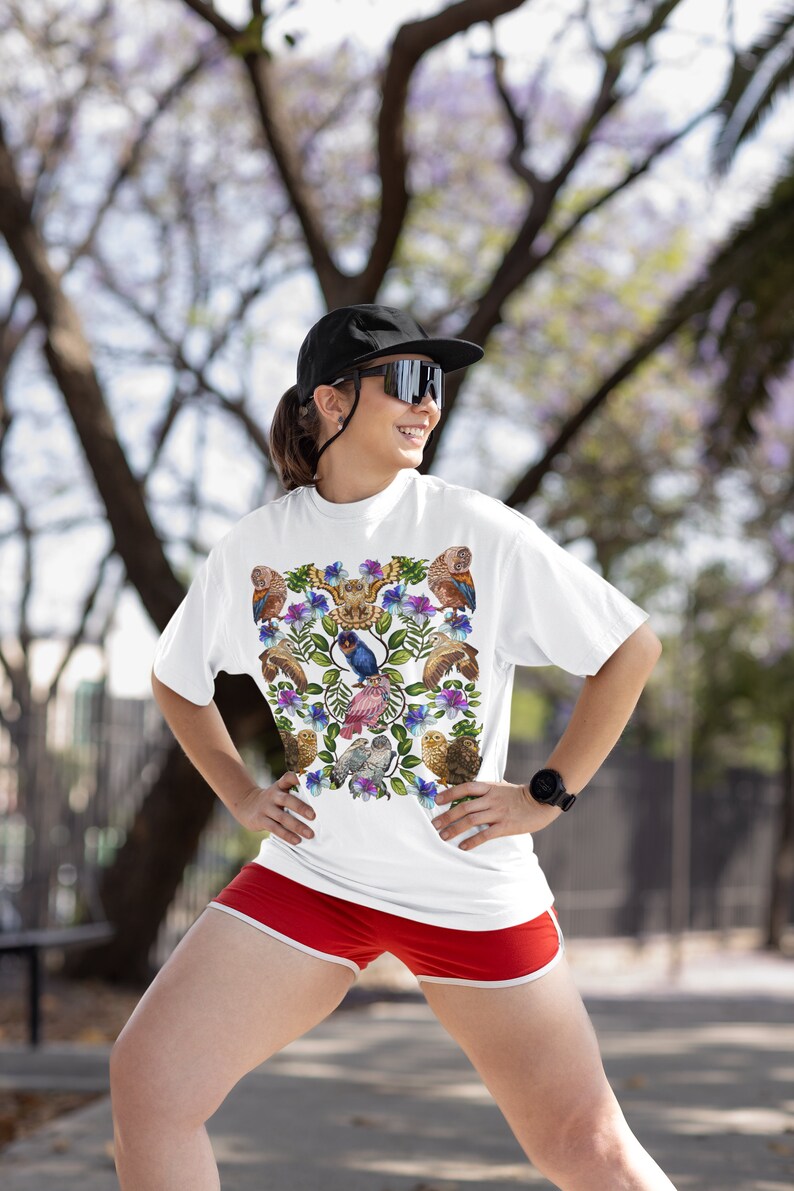 May include: White t-shirt featuring a colorful bird and floral design. The shirt has a crew neck and short sleeves. The person is wearing red shorts and a black cap and sunglasses.
