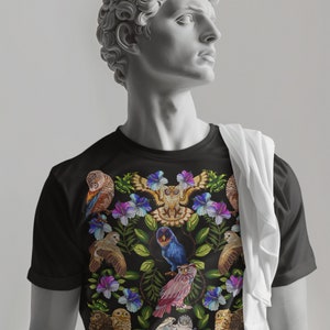 May include: Black t-shirt featuring a colorful owl and floral design. The graphic includes various owls in shades of brown, blue, and pink, surrounded by purple and blue flowers and green leaves. A white cloth is draped over the shoulder.