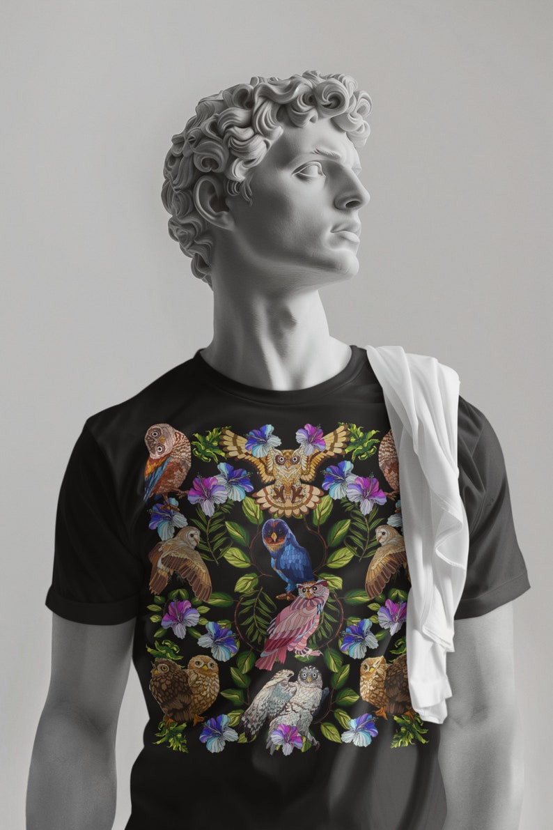 May include: Black t-shirt featuring a colorful owl and floral design. The graphic includes various owls in shades of brown, blue, and pink, surrounded by purple and blue flowers and green leaves. A white cloth is draped over the shoulder.