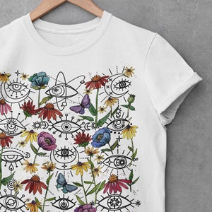 May include: White t-shirt featuring a colourful print of stylised eyes, flowers, and butterflies. The design incorporates a range of floral elements, including blue, pink, and red flowers, as well as yellow and white daisies. The eyes are drawn in a simple, geometric style and are surrounded by black lines and dots.