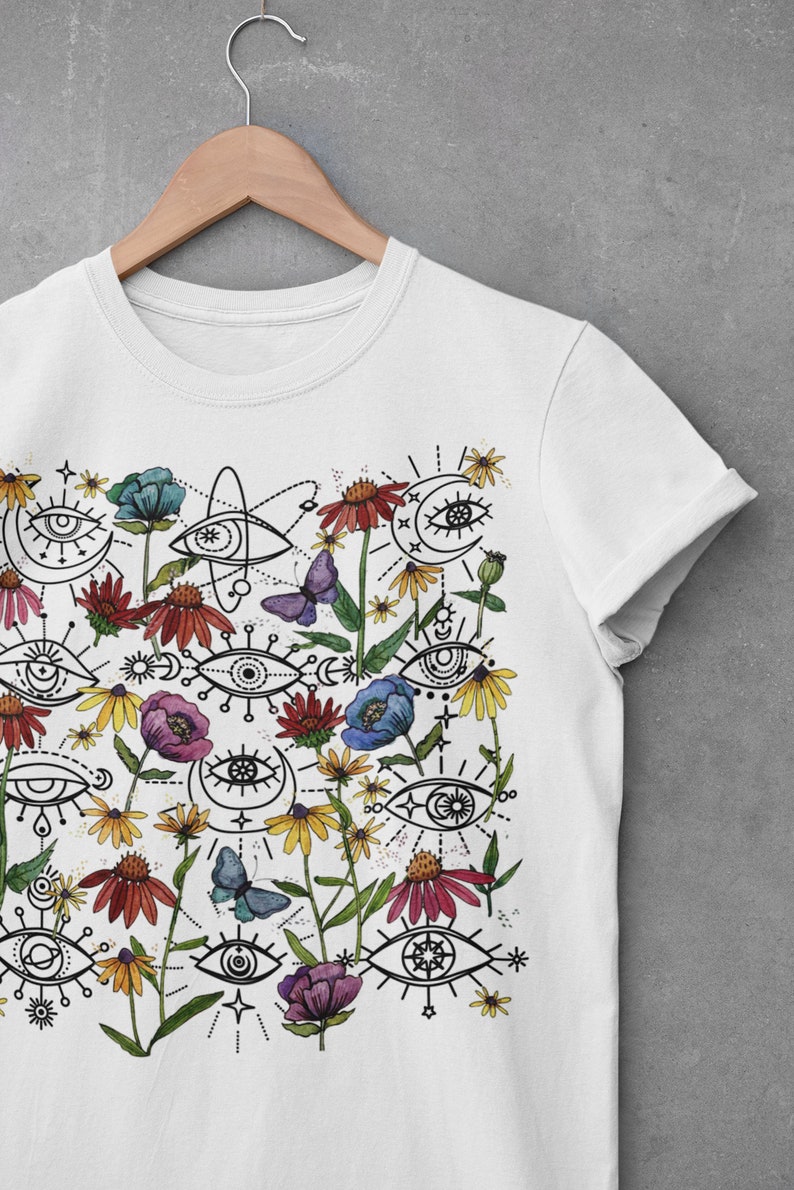 May include: White t-shirt featuring a colourful print of stylised eyes, flowers, and butterflies. The design incorporates a range of floral elements, including blue, pink, and red flowers, as well as yellow and white daisies. The eyes are drawn in a simple, geometric style and are surrounded by black lines and dots.