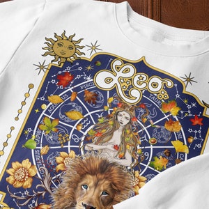 May include: White sweatshirt with a blue and gold zodiac design featuring the constellation Leo. The design includes a lion, a woman with long hair, and a circle with the zodiac signs. The text "Leo" is written in gold letters.