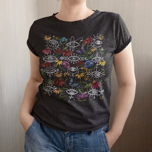 May include: A dark grey t-shirt with a white design of stylised eyes and flowers. The design features a variety of colours, including blue, green, yellow, and pink.