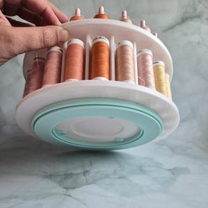 May include: A white circular rotating thread rack with two tiers holding multiple spools of sewing thread in various shades of orange, pink, and yellow. The base of the organizer is a light mint green color, and the spools are arranged vertically.