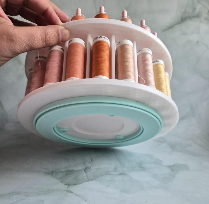 May include: A white circular rotating thread rack with two tiers holding multiple spools of sewing thread in various shades of orange, pink, and yellow. The base of the organizer is a light mint green color, and the spools are arranged vertically.