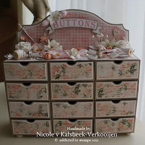 Puede incluir: Un organizador de cajones de madera rosa y blanco con la palabra "BUTTONS" en la parte superior. Los cajones están decorados con motivos florales y el organizador está lleno de botones, artículos de costura y otros artículos de artesanía.