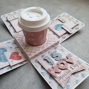 May include: A pink and blue explosion box with a coffee cup in the centre. The box has a chequered pattern and is decorated with balloons, hearts, and the words "Happy Birthday", "Special Day", "I Love You", and "15 Nov".