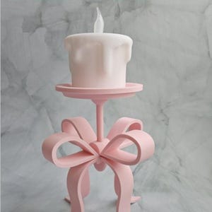 Può includere: Un portacandele rosa stampato in 3D con un design a fiocco. La candela è bianca e ha una parte superiore a forma di fiamma. Il testo in alto dice: "Questo è un file per stampante 3D. Quindi puoi farlo solo con una stampante 3D!!"