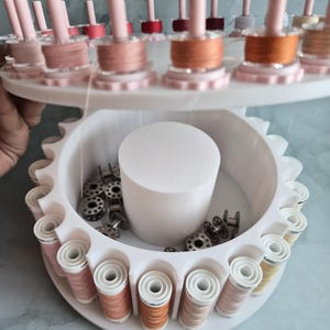 May include: A white tiered rotating thread rack featuring multiple vertical pegs holding spools of sewing thread in various shades of pink, orange, and beige. The lower section contains a central white cylinder and loose metal bobbins.