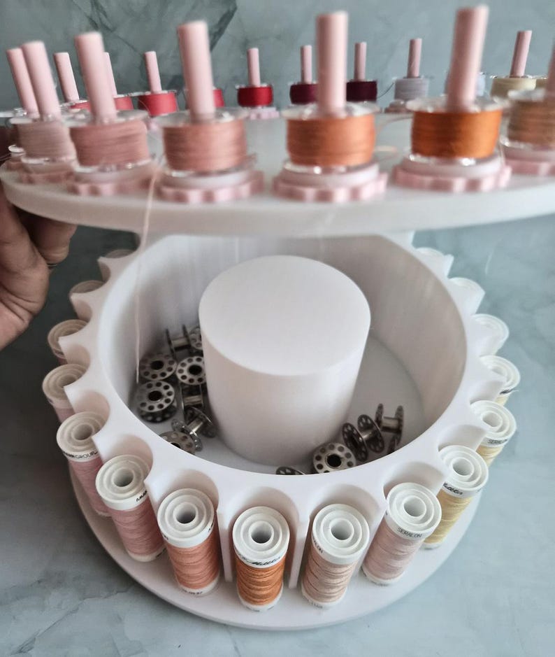 May include: A white tiered rotating thread rack featuring multiple vertical pegs holding spools of sewing thread in various shades of pink, orange, and beige. The lower section contains a central white cylinder and loose metal bobbins.