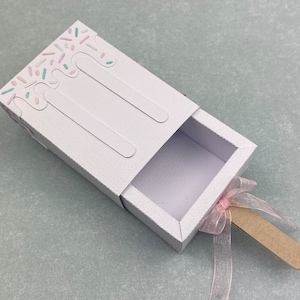 Puede incluir: Una caja de regalo blanca con forma de paleta con un cajón extraíble. La parte superior de la caja tiene un diseño de chispas y detalles de goteo. Una cinta rosa y un palito de madera están unidos a la parte inferior.