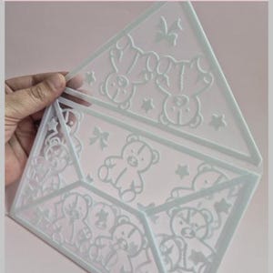 Op de afbeelding: Een wit 3D-geprint object in de vorm van een envelop met reliëfmotieven van teddyberen, sterren en strikjes. De tekst bovenaan luidt: This is a 3D Printer File. So you can only make this on a 3D Printer!! De structuur is plat en open.