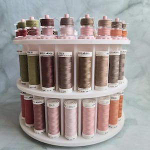 May include: A white tiered rotating thread rack holding numerous spools of Seralon and Mettler sewing thread in various shades of brown, tan, pink, and green. The spools are arranged in two circular rows on the plastic stand.