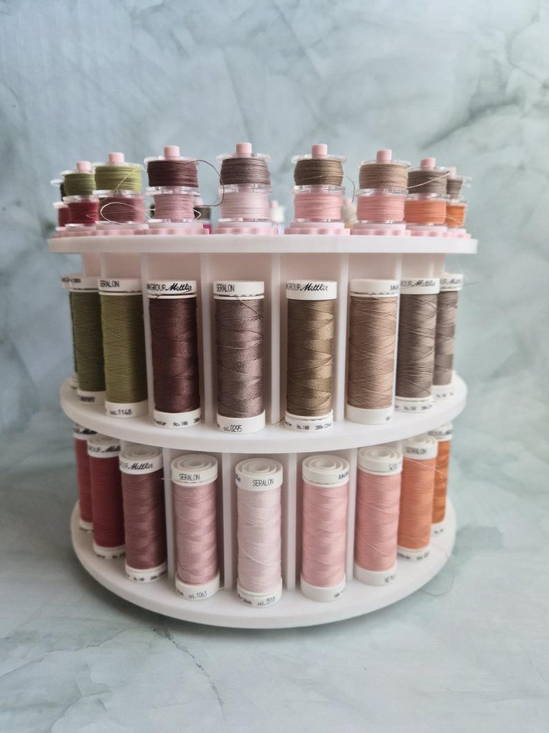 May include: A white tiered rotating thread rack holding numerous spools of Seralon and Mettler sewing thread in various shades of brown, tan, pink, and green. The spools are arranged in two circular rows on the plastic stand.
