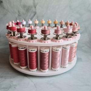 May include: A white circular rotating thread rack holding multiple spools of sewing thread in various shades of pink, ranging from deep rose to pale blush. Each spool is labeled with the text Mettler and sits in a dedicated slot.