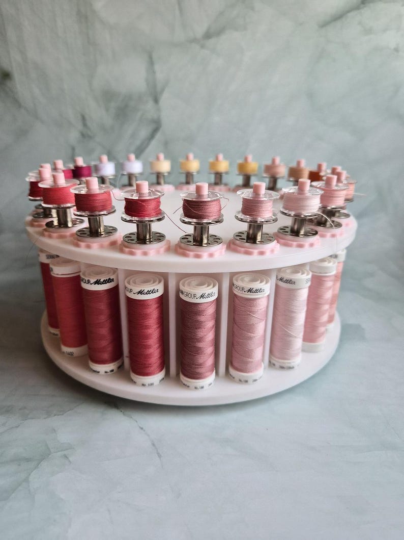 May include: A white circular rotating thread rack holding multiple spools of sewing thread in various shades of pink, ranging from deep rose to pale blush. Each spool is labeled with the text Mettler and sits in a dedicated slot.