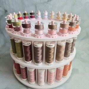 May include: A white tiered rotating thread rack holding numerous spools of Mettler Seralon sewing thread in various shades of pink, brown, green, and beige. The spools are arranged in circular rows on the plastic stand, displaying labels with brand text.