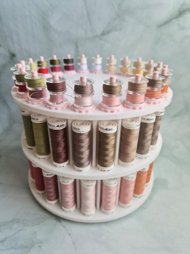 May include: A white tiered rotating thread rack holding numerous spools of Mettler Seralon sewing thread in various shades of pink, brown, green, and beige. The spools are arranged in circular rows on the plastic stand, displaying labels with brand text.