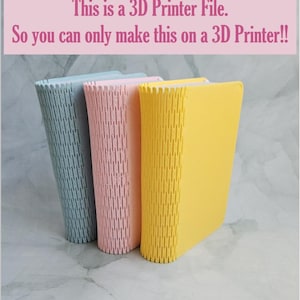 Könnte beinhalten: Drei 3D-gedruckte Bücher, eines blau, eines rosa und eines gelb. Die Bücher sind übereinander gestapelt. Der Text auf dem Bild lautet "Dies ist eine 3D-Druckdatei. Sie können dies also nur mit einem 3D-Drucker herstellen! !"