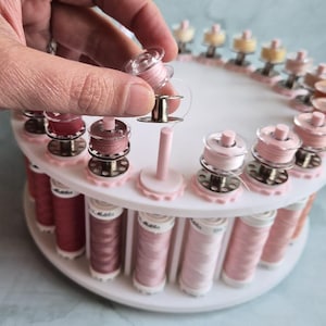 May include: A white circular sewing thread organizer stand holding multiple spools of thread in various shades of pink and rose. The stand features vertical pegs for individual bobbins, with one clear plastic bobbin being held above the base.