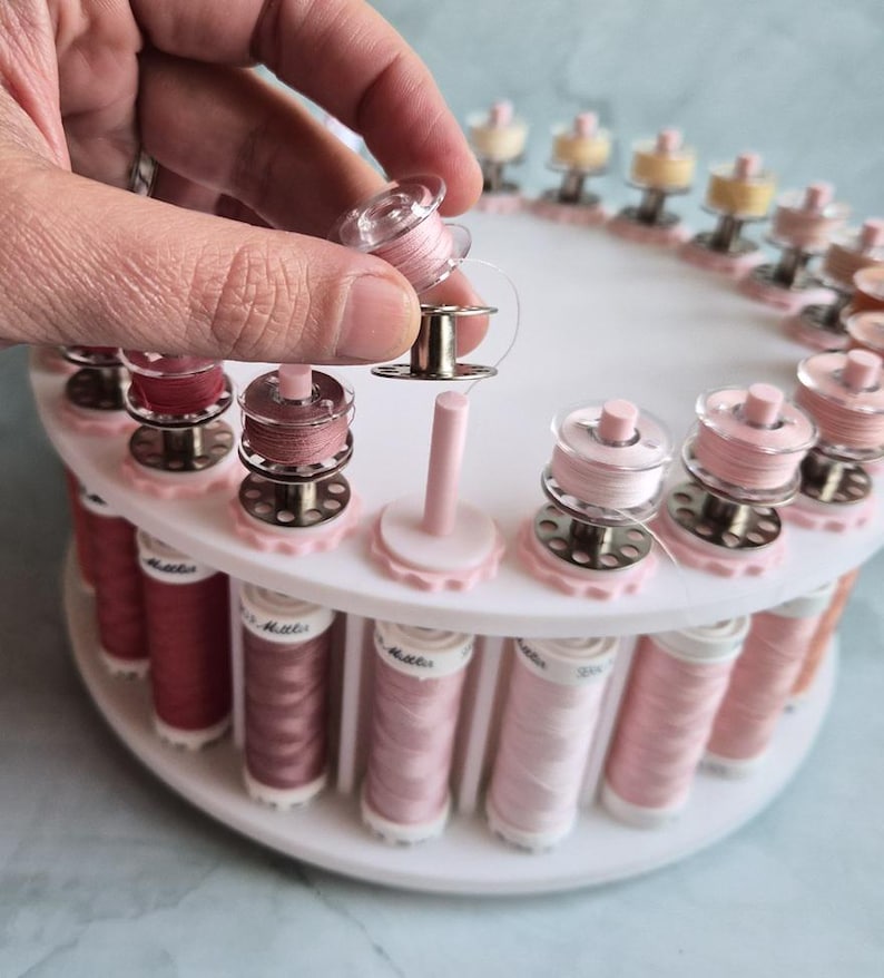 May include: A white circular sewing thread organizer stand holding multiple spools of thread in various shades of pink and rose. The stand features vertical pegs for individual bobbins, with one clear plastic bobbin being held above the base.