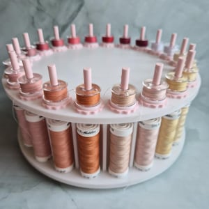 May include: A white circular rotating thread rack holding multiple spools of sewing thread in various shades of pink, orange, and yellow. Each spool is positioned on a vertical peg, with the brand name Mettler visible on the labels.