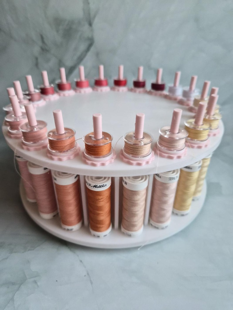 May include: A white circular rotating thread rack holding multiple spools of sewing thread in various shades of pink, orange, and yellow. Each spool is positioned on a vertical peg, with the brand name Mettler visible on the labels.