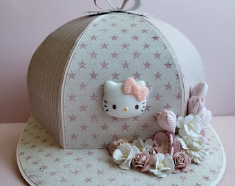 May include: A white and pink Hello Kitty character is attached to a light pink and white paper hat with a white bow. The hat has a white brim with a light pink and white star pattern. The hat is decorated with white and pink flowers.