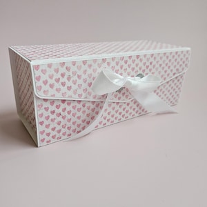 Może przedstawiać: Białe pudełko prezentowe z wzorem różowych serduszek i białą satynową wstążką zawiązaną w kokardkę.