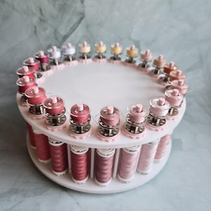 May include: A white circular tiered thread rack holding numerous spools of sewing thread in various shades of pink, red, and neutral tones. The top tier features small bobbins, while the bottom tier holds larger spools of thread arranged in a ring.