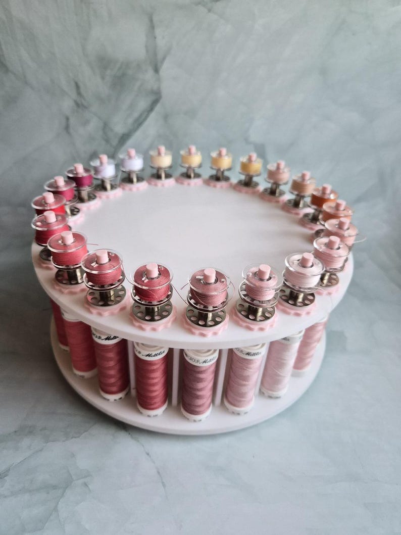 May include: A white circular tiered thread rack holding numerous spools of sewing thread in various shades of pink, red, and neutral tones. The top tier features small bobbins, while the bottom tier holds larger spools of thread arranged in a ring.