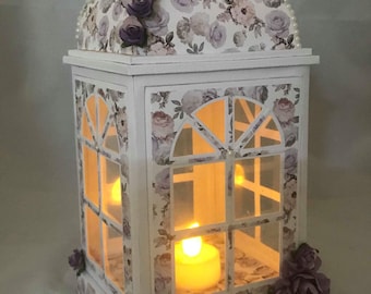 May include: A white lantern with a floral design and a purple flower garland on the top. The lantern has a clear glass window and a candle inside.