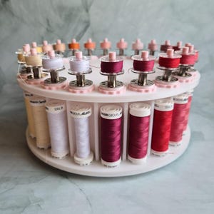 May include: A white circular rotating thread rack holding multiple spools of Amann Group Seralon polyester thread in various shades of pink, red, and cream, with matching bobbins secured on top of each corresponding spool.