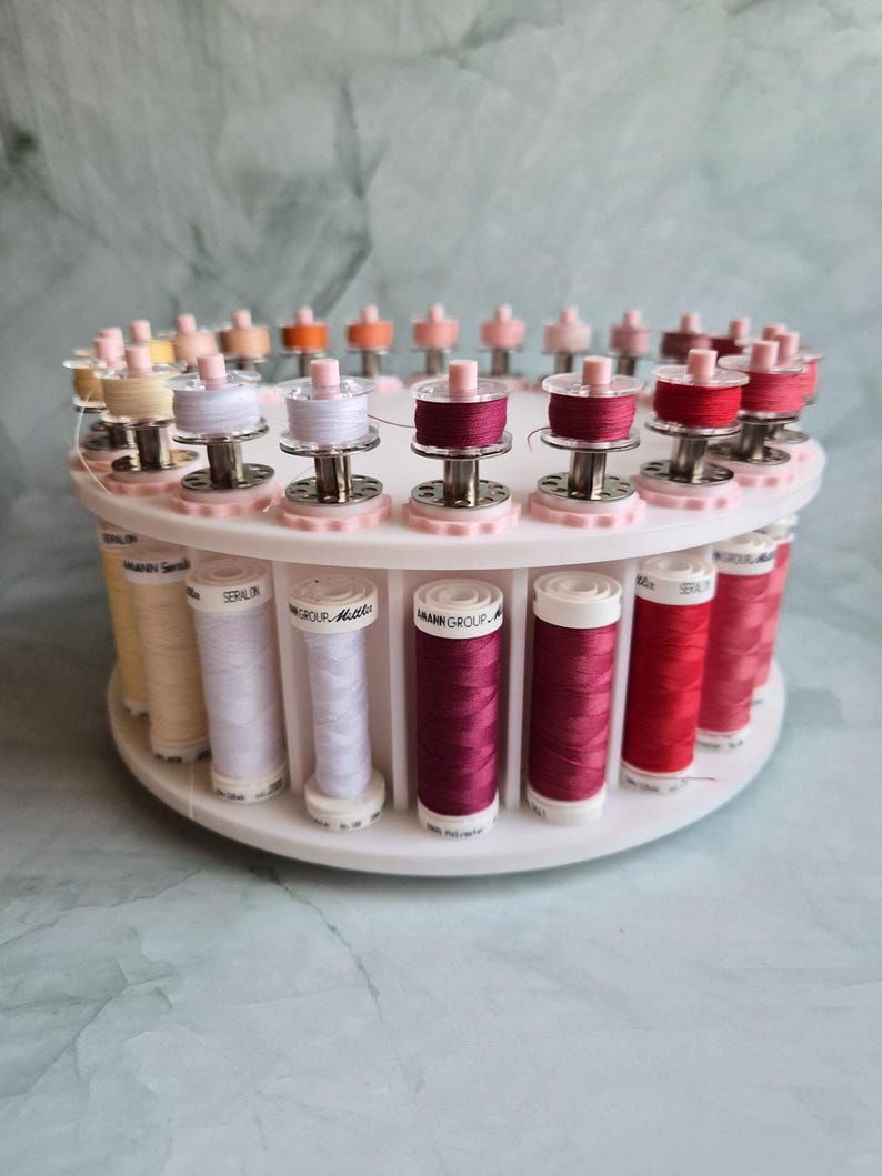 May include: A white circular rotating thread rack holding multiple spools of Amann Group Seralon polyester thread in various shades of pink, red, and cream, with matching bobbins secured on top of each corresponding spool.