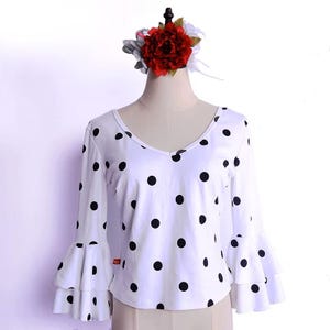 May include: White blouse with black polka dots and long sleeves with ruffled cuffs. The blouse has a V-neck and is displayed on a mannequin. A red and white flower hair accessory is on the mannequin's head.