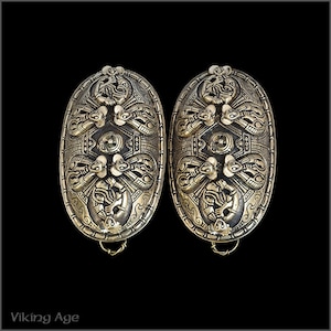 May include: Two oval-shaped, silver-toned metal brooches with intricate, detailed designs. The brooches feature a central motif of a stylized animal, surrounded by smaller animal figures and geometric patterns.