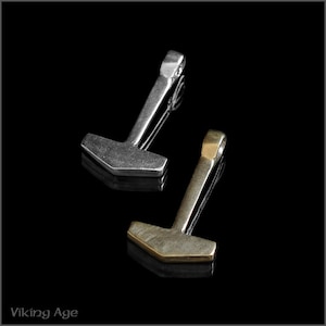 Könnte beinhalten: Zwei Metall-Hammer-Anhänger, einer silberfarben und einer golden, auf schwarzem Hintergrund. Die Anhänger sind in Form eines traditionellen Hammers mit einem abgerundeten Kopf und einem langen Griff geformt.