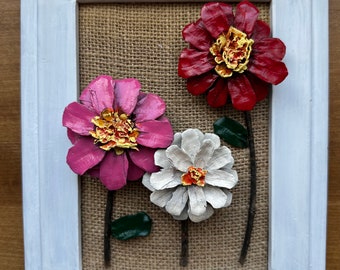 Puede incluir: Una obra de arte enmarcada con tres flores de piña pintadas en rosa, blanco y rojo. Las flores están dispuestas sobre un fondo de arpillera y tienen tallos de rama.