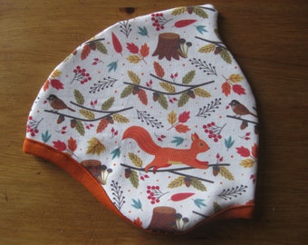 Puede incluir: Un gorro de tela blanca con ribete naranja. El gorro tiene un patrón de bosque con ardillas, pájaros, hojas y bellotas.