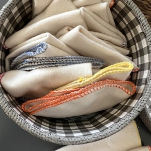 May include: A basket filled with white cotton dishcloths with colourful embroidered edges. The basket has a black and white checked pattern.