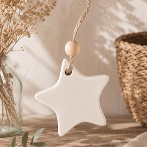 Peut inclure: Une étoile en céramique blanc cassé à la surface lisse, suspendue par une ficelle en jute naturelle. Une perle en bois clair de forme ronde est enfilée sur la ficelle juste au-dessus de l'étoile, qui comporte un petit trou pour la suspension.